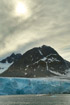 The blue-white front of a glacier reaches the sea over a boulder beach in Spitzbergen with a dim sun showing through overcast