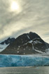 The blue-white front of a glacier reaches the sea over a boulder beach in Spitzbergen with a dim sun showing through overcast